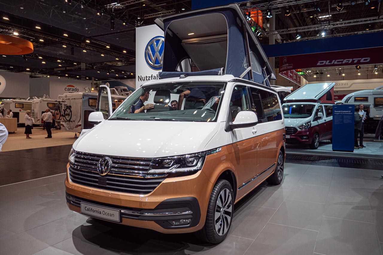 VW California 6.1 – Strand-Hopping mit Stil (inkl. Video-Roomtour) 19 Blick von vorn auf einen VW California 6.1 Ocean mit offener Heckklappe und aufgestelltem Hochdach auf dem Messestand des Caravan Salon 2019 in Düsseldorf
