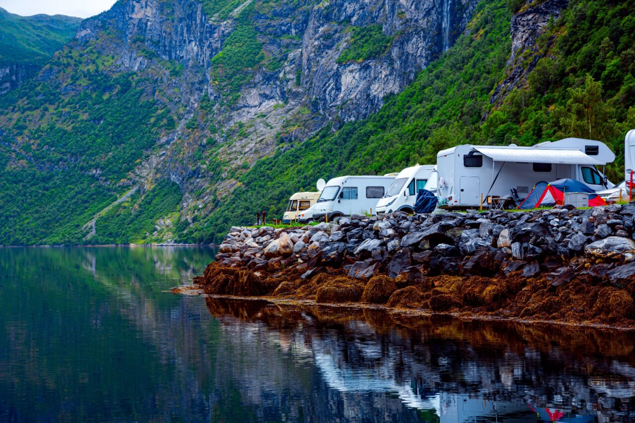 An einem See zwischen Bergen stehen verschiedene Campingfahrzeuge und ein Zelt.