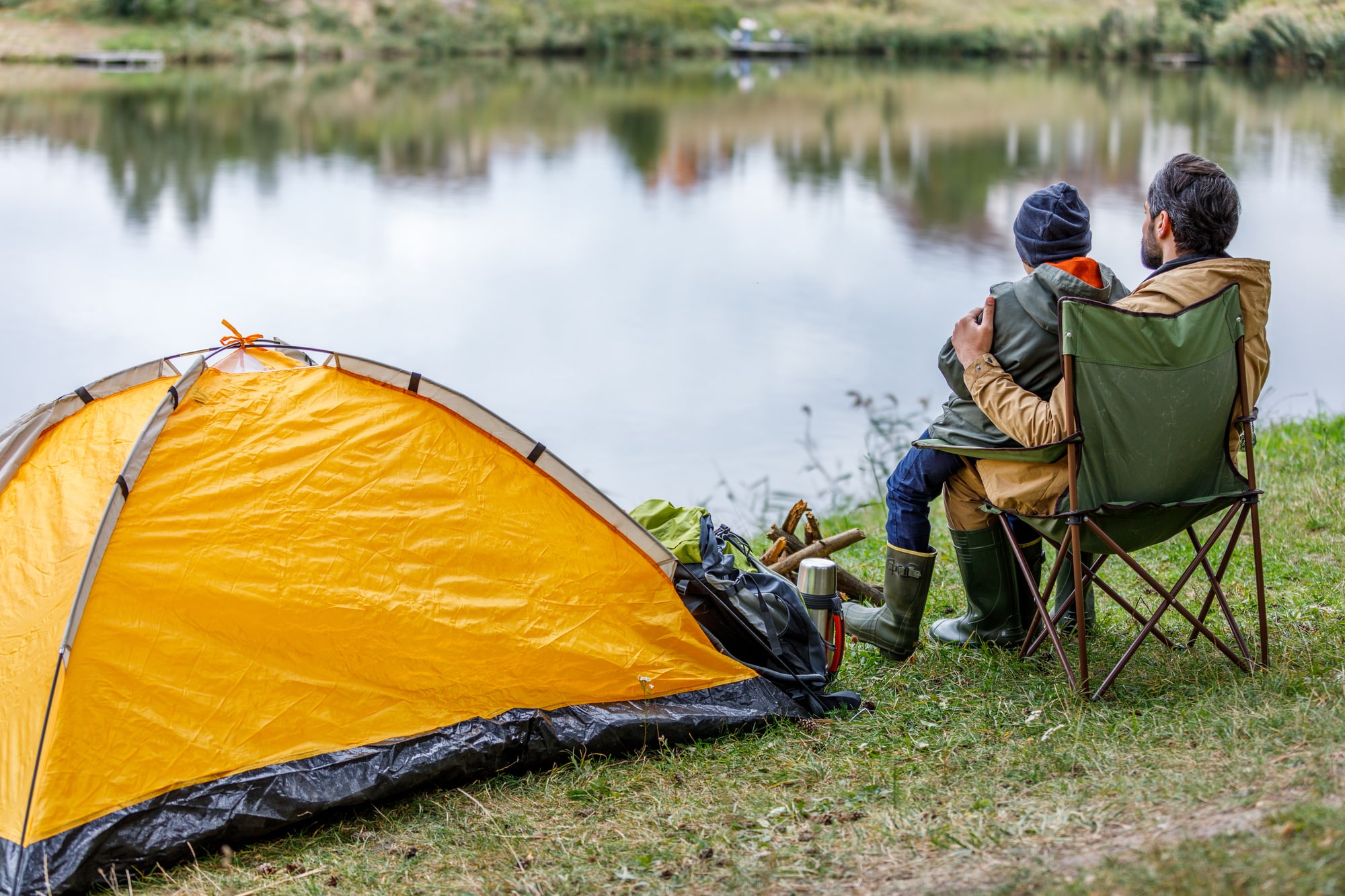 Vater und Kind sitzen am See in einem Campingstuhl neben einem Zelt