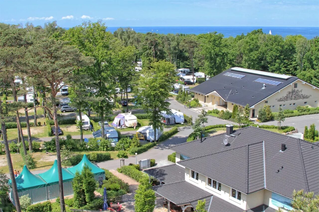 Evalué par les campeurs : Ce sont les meilleurs campings d'Europe 1 Camping.info n°1 en Allemagne : Camping Kühlungsborn. On peut voir une vue aérienne d'un camping dans la verdure, avec la mer en arrière-plan.