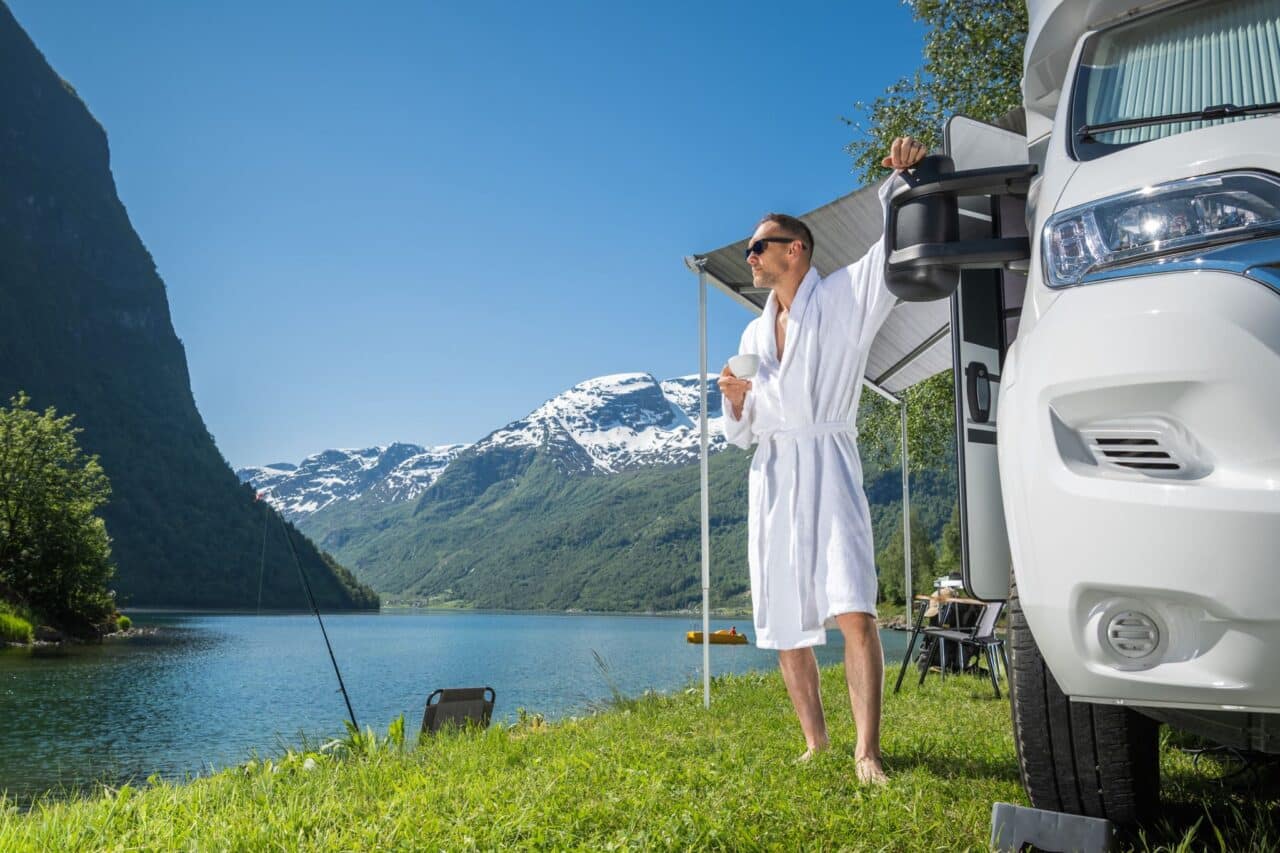 Mann im Bademantel steht mit Kaffeetasse neben Wohnmobil am See